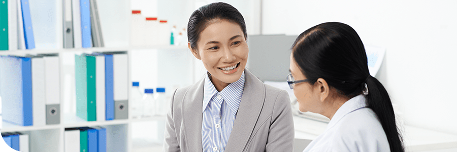 Two Asian women having a friendly conversation in an office setting about Online MHA Programs, one in a grey suit and the other in a white lab coat.