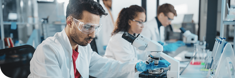 A diverse team of scientists, including a man using a microscope, working in a laboratory setting. They are focused on conducting experiments, wearing lab coats and protective eyewear.