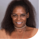 A portrait of a smiling woman with shoulder-length curly hair, wearing a light peach top and a delicate necklace. She has glasses resting on top of her head.
