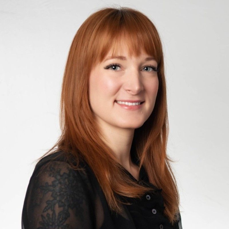 A professional headshot of a smiling woman with long red hair and a black lace-detailed blouse against a light gray backdrop.