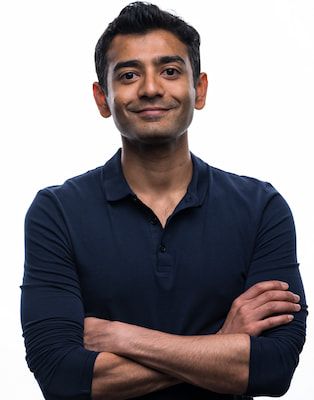 A confident man with dark hair, wearing a blue polo shirt, stands with his arms crossed, smiling slightly at the camera against a white background.
