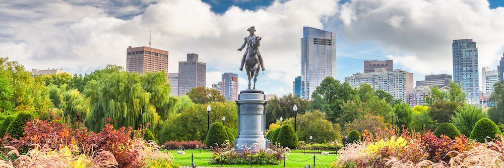 Panoramic view of a vibrant public park featuring a prominent statue of a historical figure on a pedestal, surrounded by colorful floral arrangements, with a backdrop of modern city skyscrapers under a cloudy blue sky.