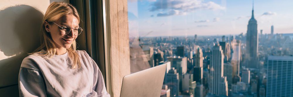 A woman wearing glasses smiles while using a laptop, seated beside a window overlooking a cityscape with skyscrapers during a sunny day.
