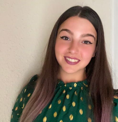 A young woman with long brown hair, wearing a green dress with yellow patterns, smiles warmly at the camera, her makeup subtly enhancing her features.