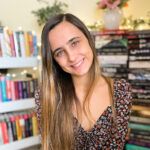 A smiling young woman with long brown hair, wearing a floral dress, sits in front of bookshelves filled with colorful books, with twinkling lights and a vase of flowers in the background. She is an enthusiastic graduate student focusing on educational strategies for individuals with ADHD.