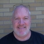 A smiling bald man with a beard, wearing glasses on his head and a black T-shirt, standing against a brick wall background.