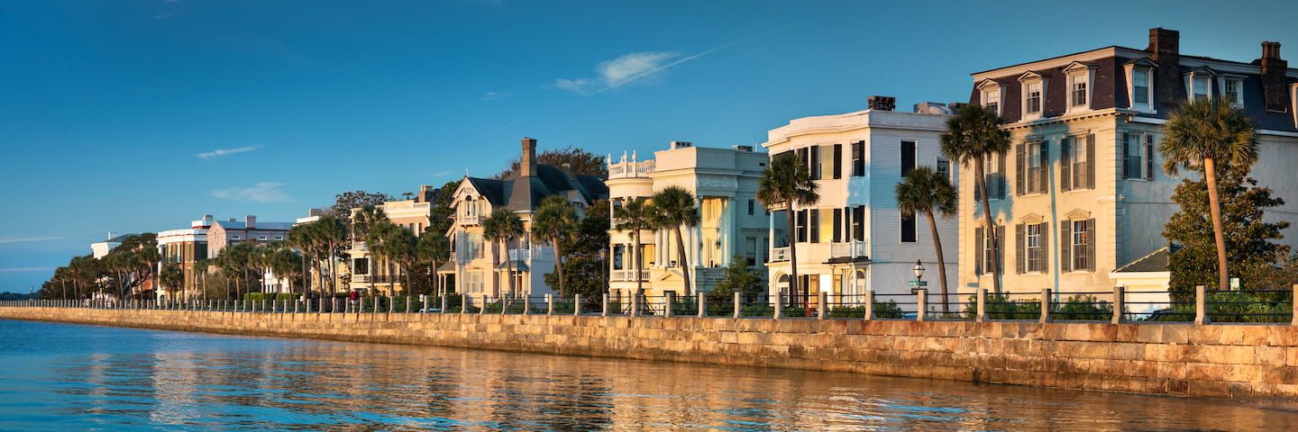 A panoramic view of a row of elegant, multistory houses along a waterfront, with calm waters in the foreground and a clear blue sky above.