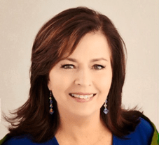 A portrait of a smiling woman with shoulder-length brown hair wearing a blue blouse and decorative earrings, set against a light neutral background.