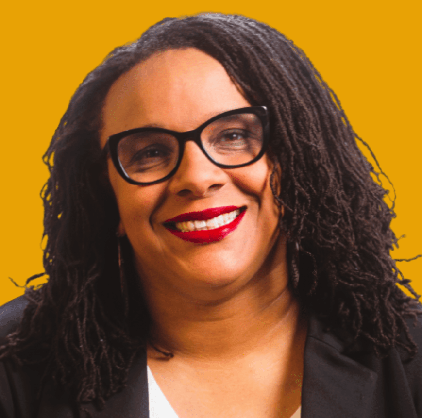 A smiling woman with shoulder-length curly hair, wearing glasses and a black blazer, against a bright yellow background. She has red lipstick on.