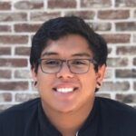 A smiling young man with glasses, wearing a black shirt, in front of a brick wall background.