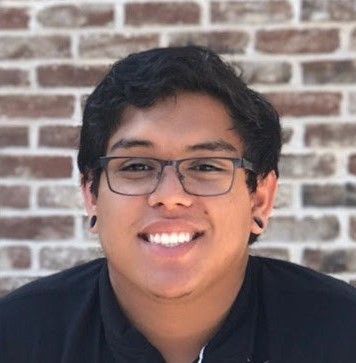 A smiling young man with glasses, wearing a black shirt, in front of a brick wall background.