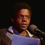 A man with glasses, wearing a sweater over a striped shirt, speaks into a microphone while reading from a paper in a dimly lit setting.
