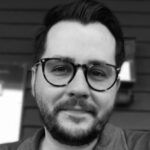Black and white portrait of a smiling man with a beard and glasses. He is facing the camera, outdoors with a blurred background, representing one of the best online MBA programs in 2023.