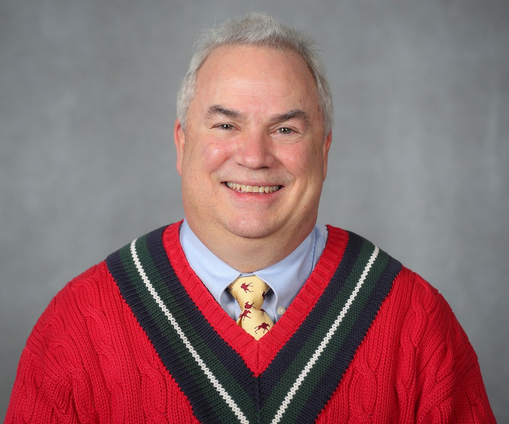 Senior man smiling, wearing a red and gray v-neck sweater over a shirt with a patterned tie. Gray background.