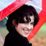 A woman with dark hair and a bright smile looking over her shoulder under a red transparent umbrella, with a blurred green background.
