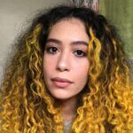 A close-up portrait of a young woman with long, curly dyed yellow hair. She has clear skin, subtle makeup, and is gazing directly at the camera.