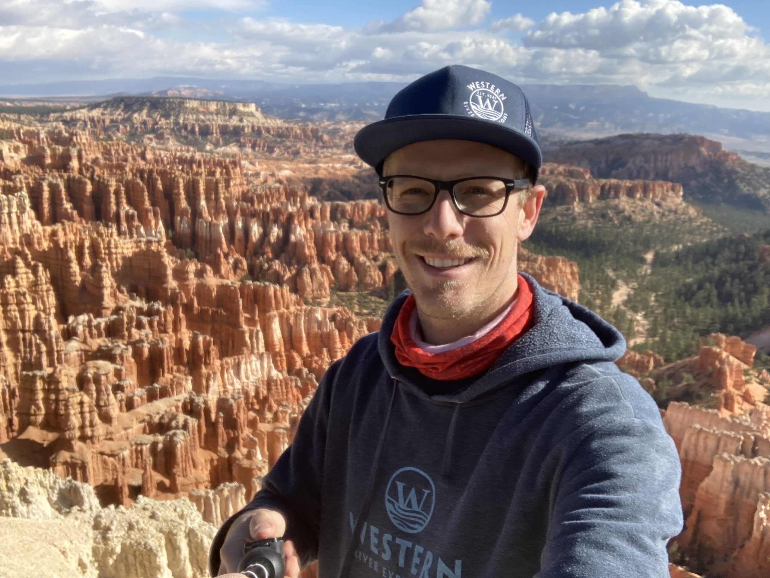 A man wearing a hat, glasses, and a sweatshirt takes a selfie with the scenic backdrop of a rugged canyon with red rock formations under a partly cloudy sky as part of developing digital literacy.
