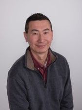 A portrait of a smiling man wearing a gray sweater over a red shirt against a light gray background.