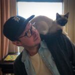 A man in glasses and a baseball cap with a Siamese cat perched on his shoulder, both looking in the same direction, in a warmly lit room.