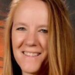 A close-up portrait of a smiling woman with long blonde hair, featuring a warm, blurred background in earthy tones.