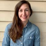 A smiling woman with long brown hair wearing a denim shirt, sitting in front of a beige siding wall.