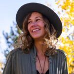 A smiling young woman wearing a black hat and olive green jacket stands outdoors, with sunlit yellow autumn leaves in the background.