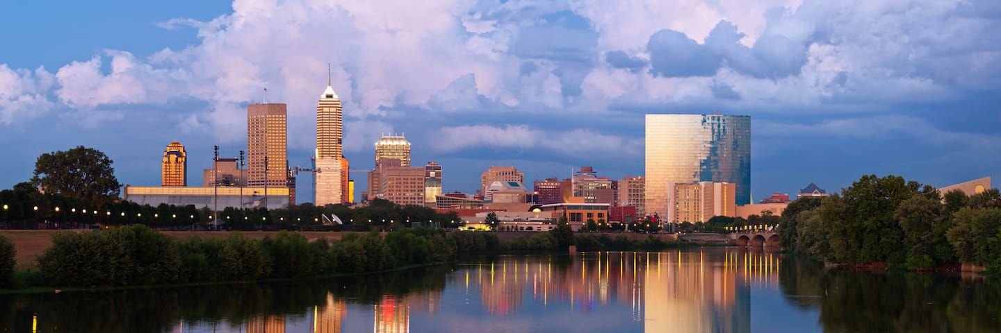 Panoramic view of an Indiana city skyline at dusk with reflections of tall buildings in a tranquil river, under a dramatic cloudy sky.