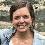 A smiling woman with brunette hair wearing a denim jacket and a pearl necklace stands in front of a stone wall.