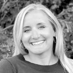 Black and white portrait of a smiling woman with blonde hair, wearing hoop earrings and a dark sweater, against a blurred foliage background.