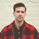 A man with short dark hair, wearing a red and black checkered shirt over a black top, stands in front of a pale brick wall, looking directly at the camera.