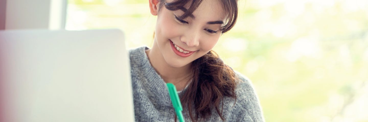 A young woman smiling while looking at her laptop screen, holding a pen and sitting in a brightly lit room.