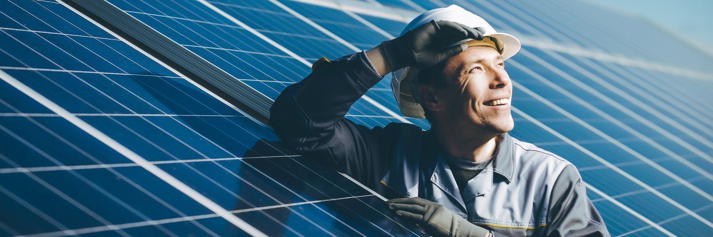 A technician in a hard hat and gloves smiles optimistically while inspecting a large solar panel array, symbolizing sustainable energy solutions.