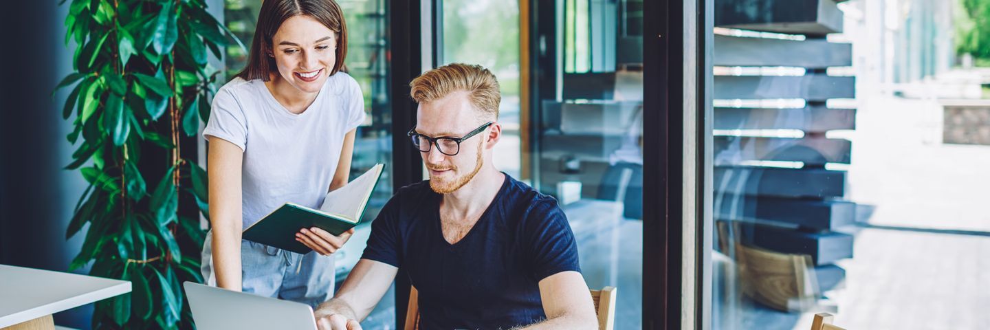 Two colleagues discussing over a notebook at a modern workspace, with the woman standing and smiling down at the seated man who is writing notes.