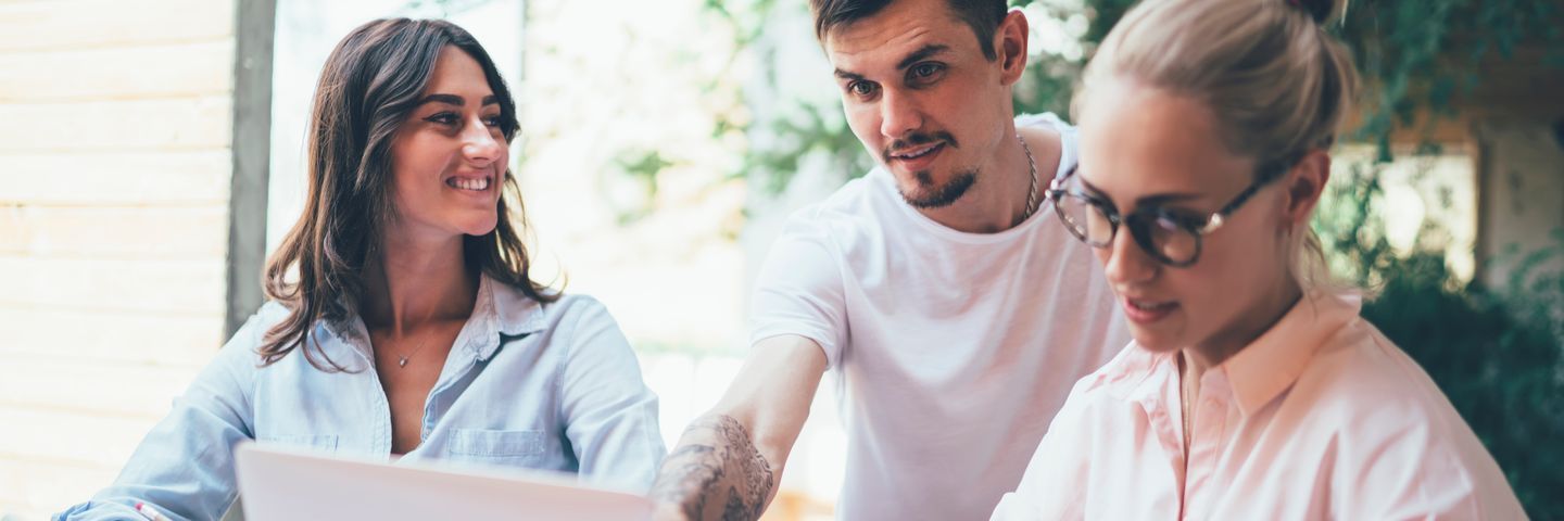 Three young adults, one man and two women, collaborate over a laptop in a bright, casual setting. they are engaged in a discussion, looking focused and interested.