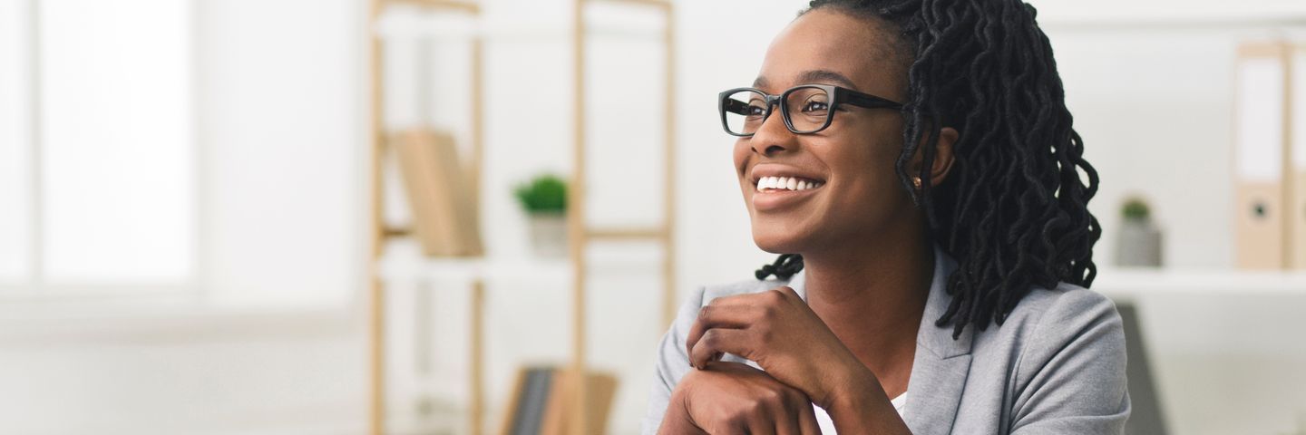 A joyful young black woman with glasses and braided hair, smiling and looking away, sitting in a bright office environment.