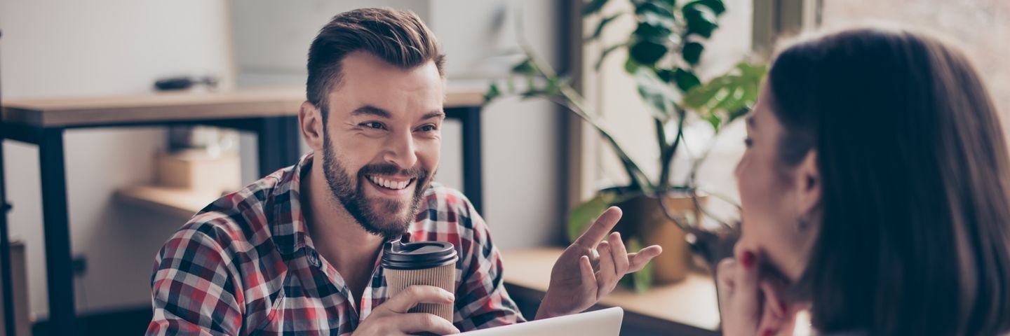 A smiling man in a plaid shirt holding a coffee cup while talking to a woman during a casual meeting. they sit at a table in a brightly lit room with plants in the background.