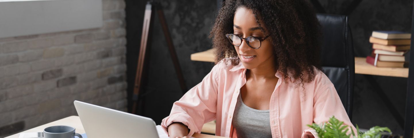 A young woman with curly hair, wearing glasses and a pink shirt, works intently on a laptop in a modern office setting with books and a coffee cup nearby.