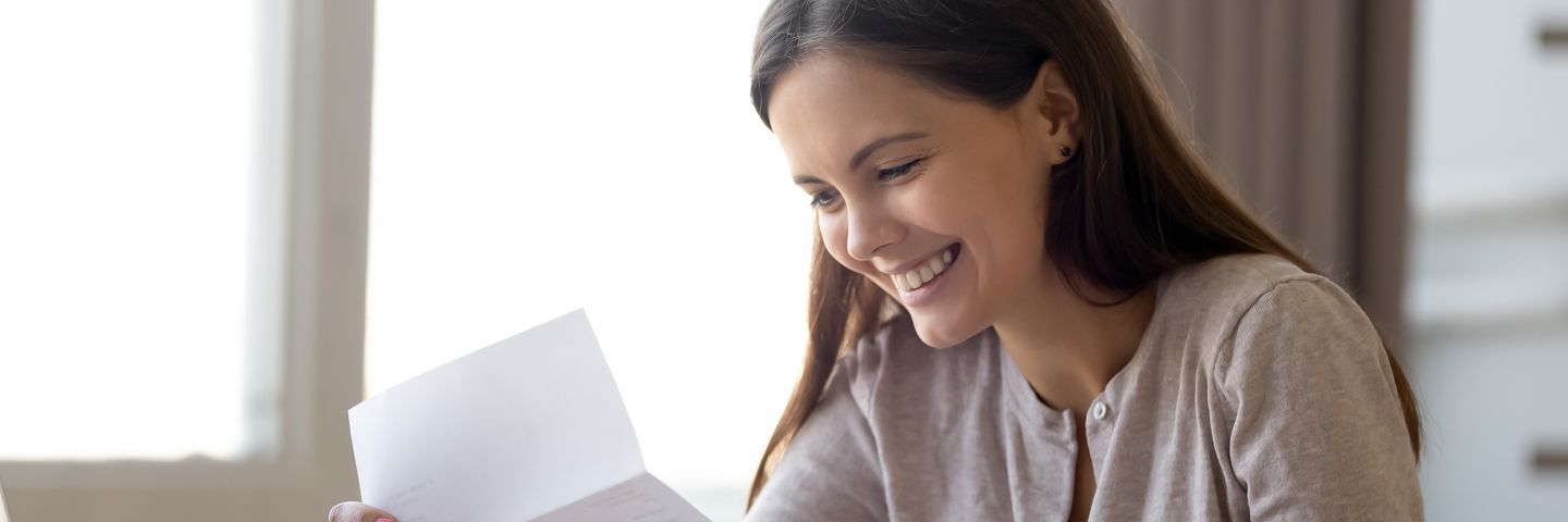 A joyful young woman reading a letter at a bright indoor setting, smiling widely with apparent happiness or good news.