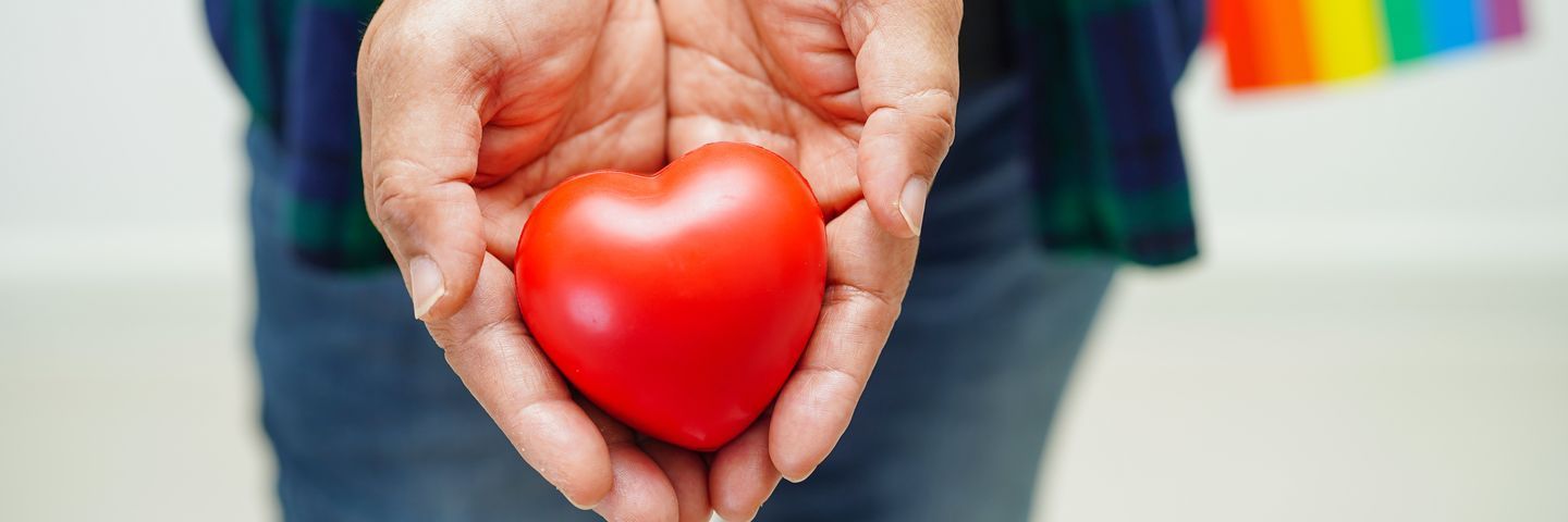 Close-up of a person holding a red heart-shaped object in their palms, with a blurred background showing rainbow colors.