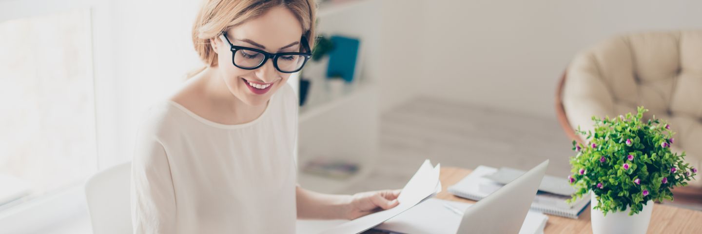 A smiling woman with glasses reviews documents at a bright home office desk, with a plant nearby, conveying a cheerful work environment.