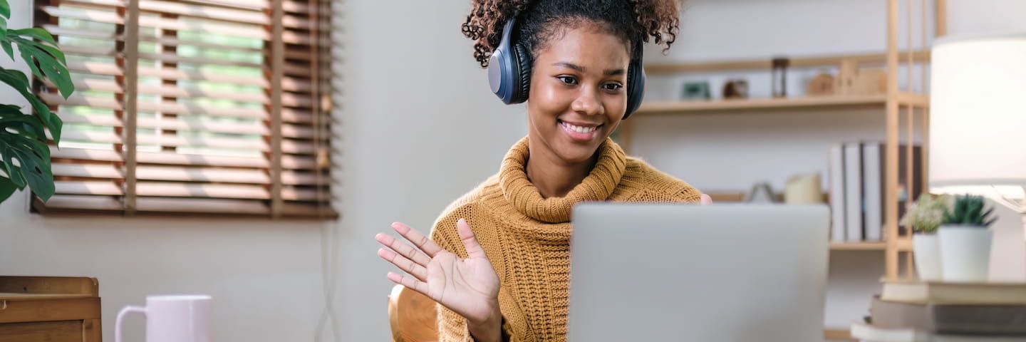 A young woman in a mustard sweater and headphones smiles while waving at her laptop screen in a bright, plant-decorated room, suggesting a friendly video call.