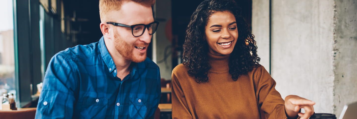 Two individuals in a casual setting. on the left, a man with ginger hair wearing glasses and a blue shirt is smiling. on the right, a woman with curly hair in a brown sweater is laughing and pointing at a laptop.