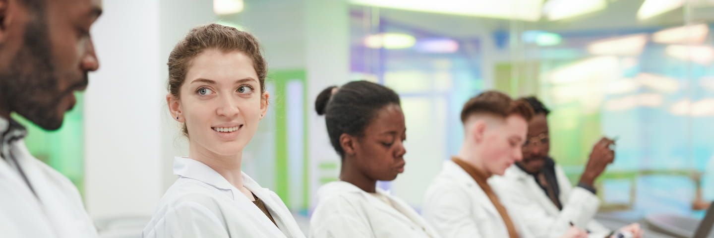 A diverse group of medical professionals seated in a row, with a young caucasian female doctor smiling at the camera, colleagues of various ethnicities focused on their notes.