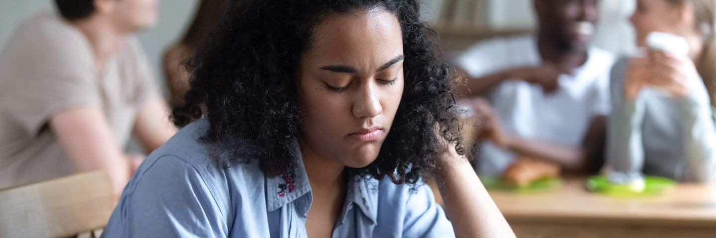 A young woman with curly hair rests her head on her hand, looking down thoughtfully, with other individuals sitting at a table in the background.