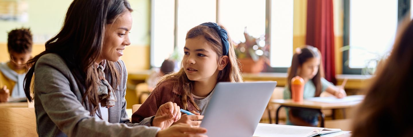 A teacher and a young student engage in a discussion over a laptop in a brightly lit classroom with other students in the background.