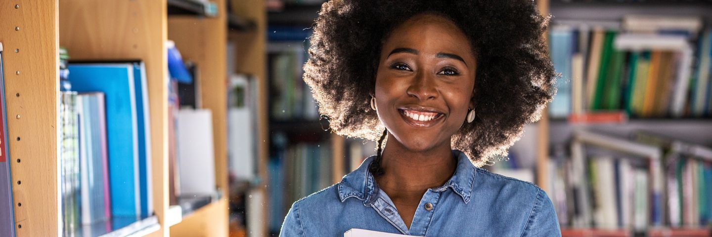 A cheerful young black woman with a voluminous afro hairstyle holding books in a library, smiling broadly at the camera.
