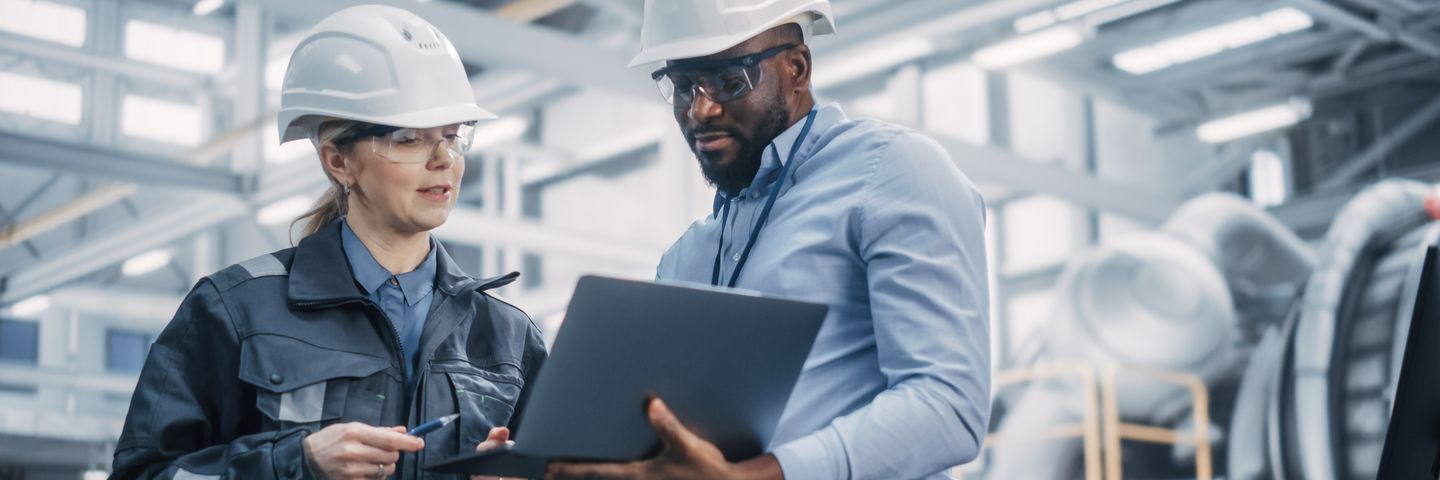 Two engineers, one female in a protective helmet and one male with glasses and a beard, discuss work on a laptop in an industrial setting.