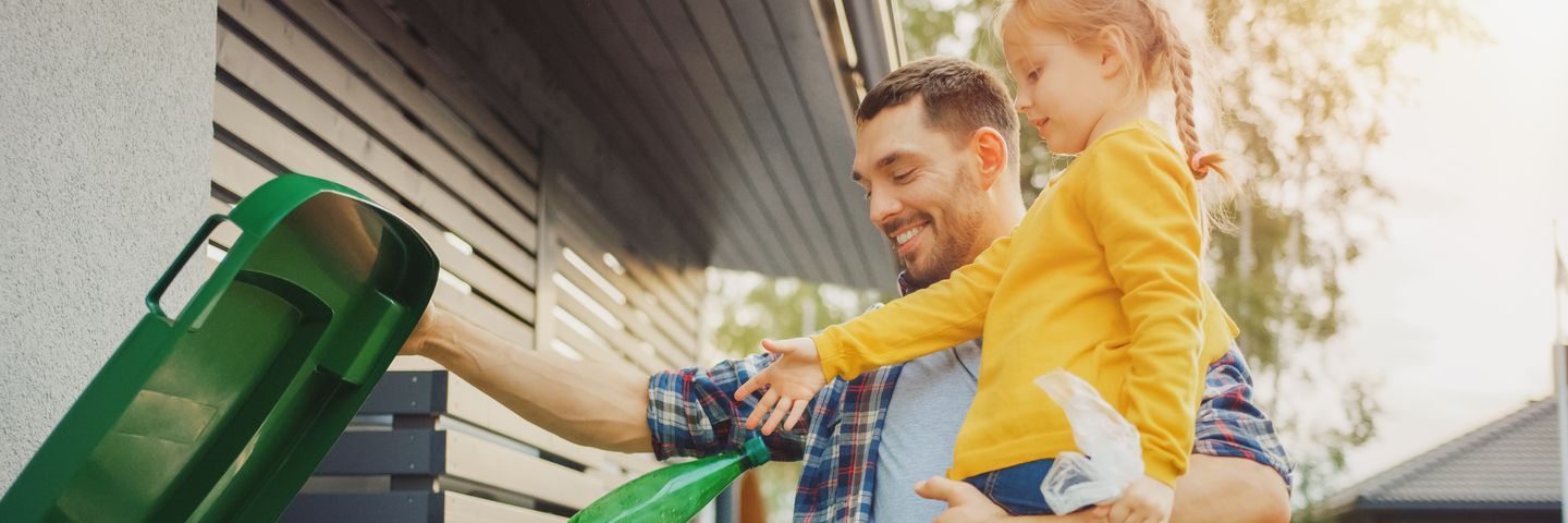 A father, smiling, helps his young daughter put a plastic bottle into a green recycling bin outside their home, promoting environmental responsibility.