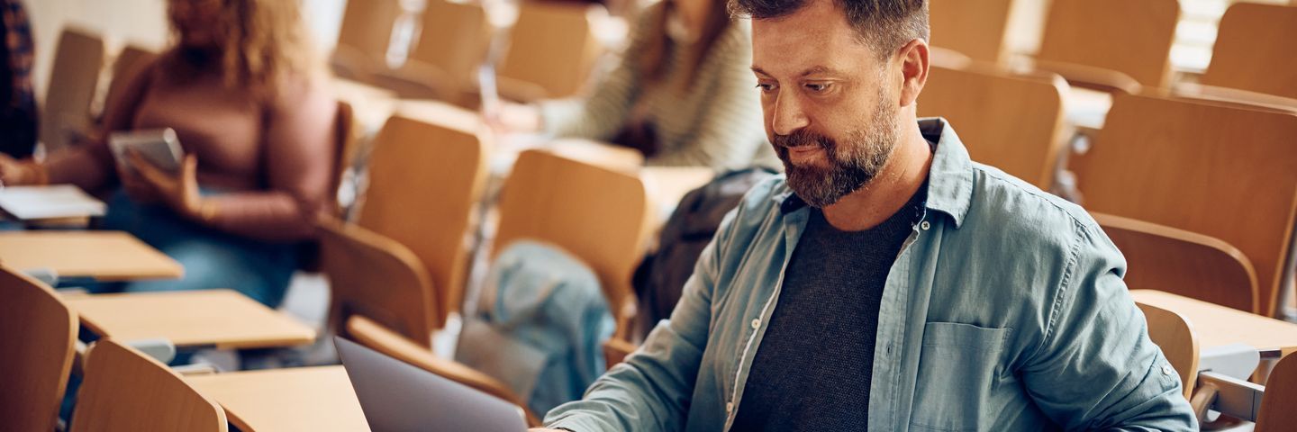 A mature male student with a beard, wearing a denim shirt, focuses on his laptop in a lecture hall filled with other students.