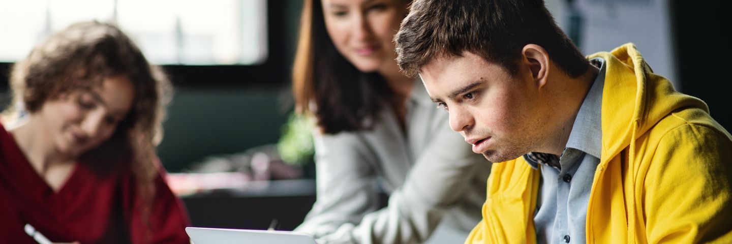 A young man in a yellow jacket intently looks at a tablet screen, sitting alongside two female colleagues who are engaged in their own work. they are in a busy, well-lit office setting.